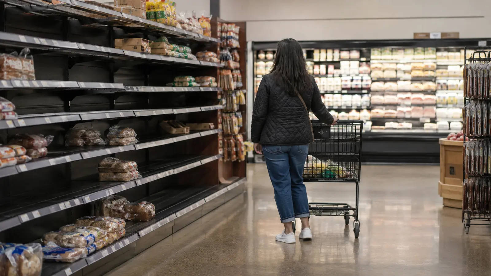 Kısmen boş market rafları arasında alışveriş arabasıyla ilerleyen bir tüketici